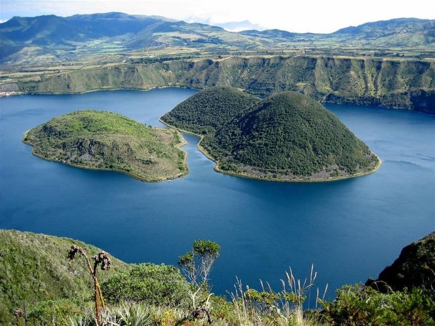 Vue plongeante de deux îles vertes dans un lac de cratère bleu profond, entouré de collines ondulantes sous un ciel clair.
