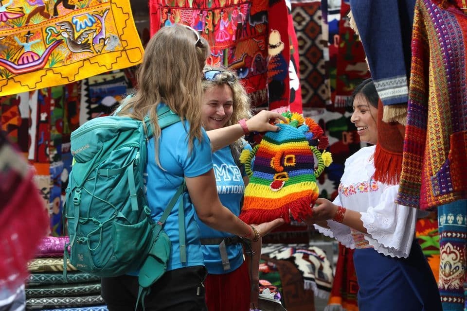 Zwei Frauen auf einer WeRoad-Gruppenreise betrachten einen farbenfrohen Strickartikel, den ein lokaler Händler auf einem Freiluftmarkt anbietet.