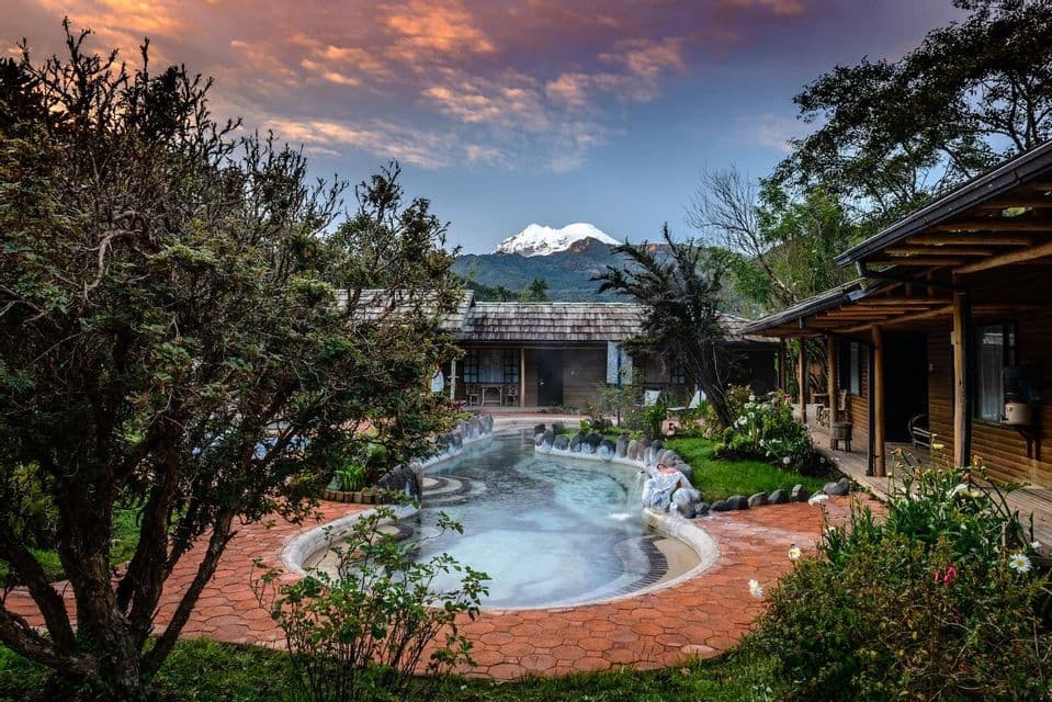 A steaming thermal pool at a resort with wooden lodges, set against a backdrop of a snow-capped mountain under a sunset sky.