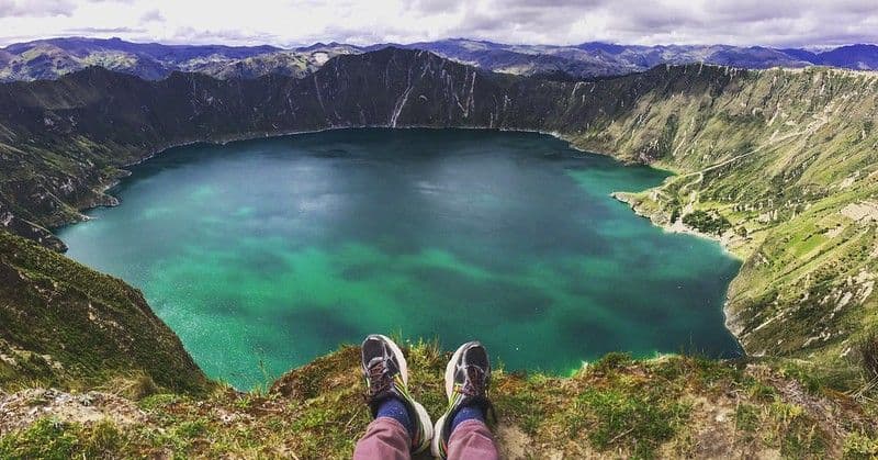 Vista en primera persona de unas piernas y botas de senderismo colgando sobre el borde de un acantilado herboso, con vistas a un lago de cráter turquesa.