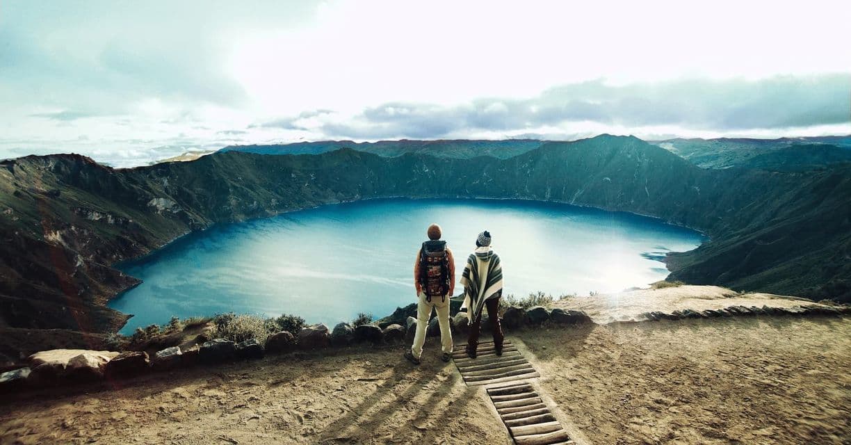 Two people seen from behind, one wearing a backpack and the other a poncho, stand at a viewpoint overlooking a crater lake.