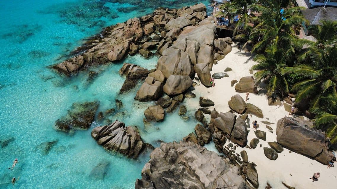 Veduta aerea di una costa rocciosa con una spiaggia di sabbia bianca, palme e persone che nuotano in acque cristalline turchesi.