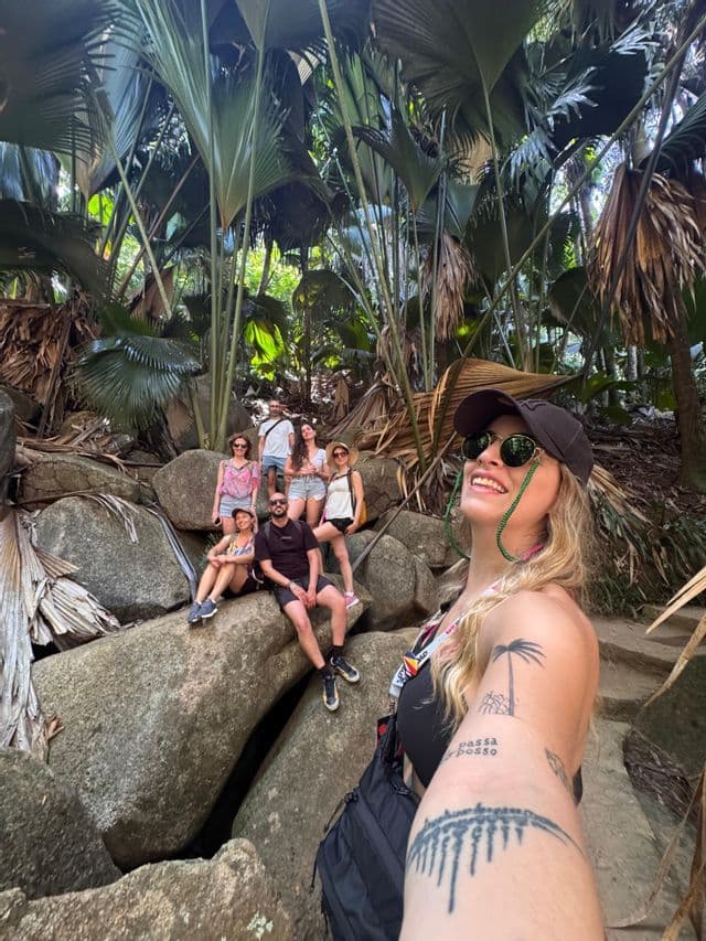 Una donna tatuata si scatta un selfie sorridente con un gruppo WeRoad in posa su grandi rocce in una lussureggiante giungla tropicale.