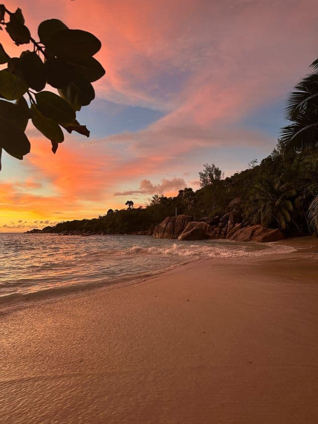 Onde lambiscono una spiaggia sabbiosa, adiacente a una costa rocciosa, sotto un cielo al tramonto vivido, arancione e rosa.