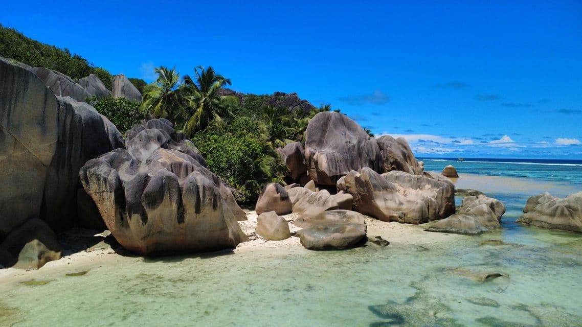 Grandi massi di granito levigati riposano su una spiaggia sabbiosa, accarezzata da acque turchesi limpide e poco profonde, con palme sullo sfondo.