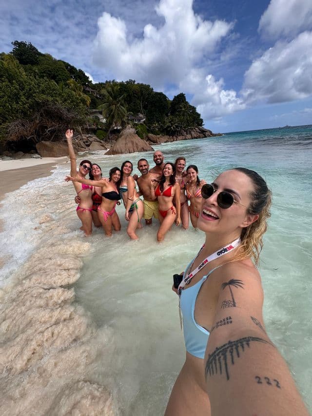 Una donna sorridente si scatta un selfie con un gruppo WeRoad, in piedi nell'acqua bassa di una spiaggia tropicale.