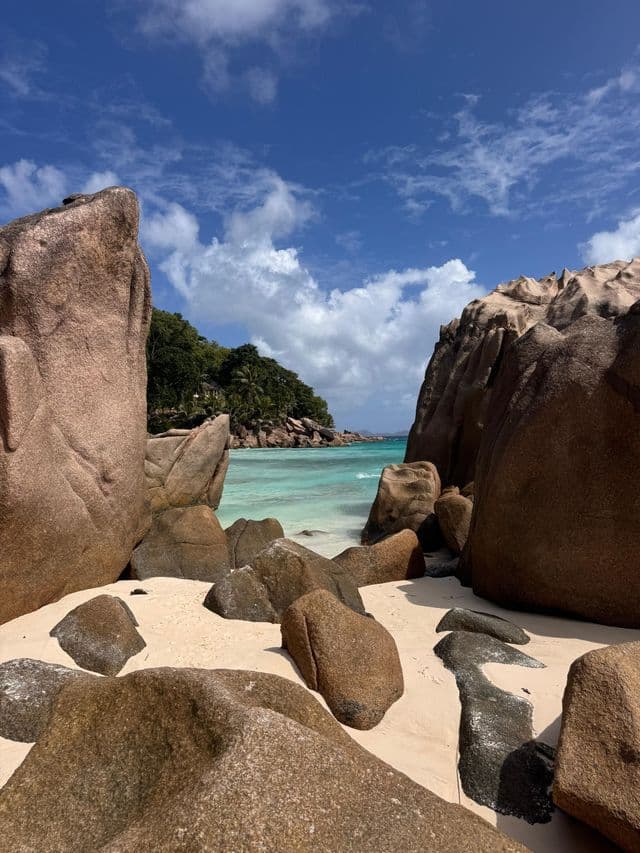 Grandi massi di granito su una spiaggia di sabbia bianca incorniciano una vista sull'oceano turchese e una costa verde e collinare.