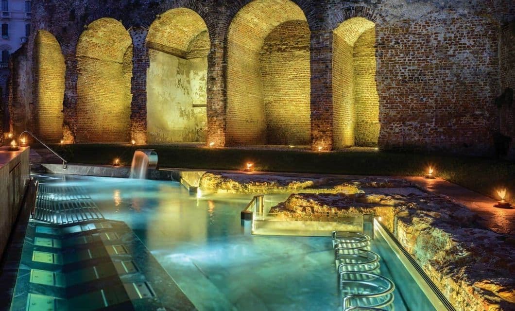 Una piscina termale all'aperto illuminata da luci calde e fiaccole di notte, con antichi archi in mattoni sullo sfondo.