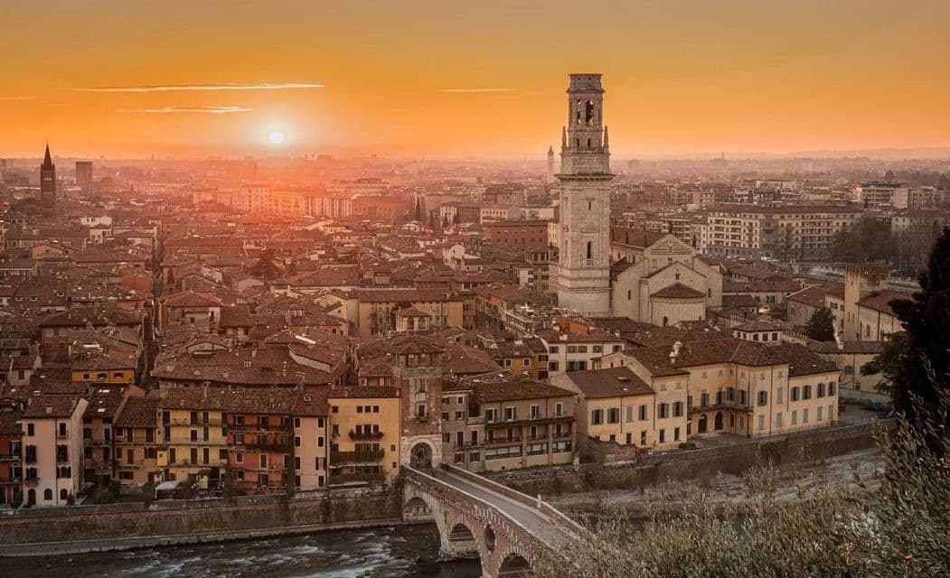 Una veduta aerea di una città storica con un ponte di pietra e un campanile sotto un tramonto arancione splendente.