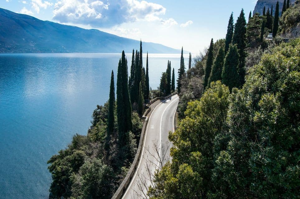 Vista aerea di una strada tortuosa lungo una scogliera accanto a un grande lago blu, con montagne sullo sfondo.