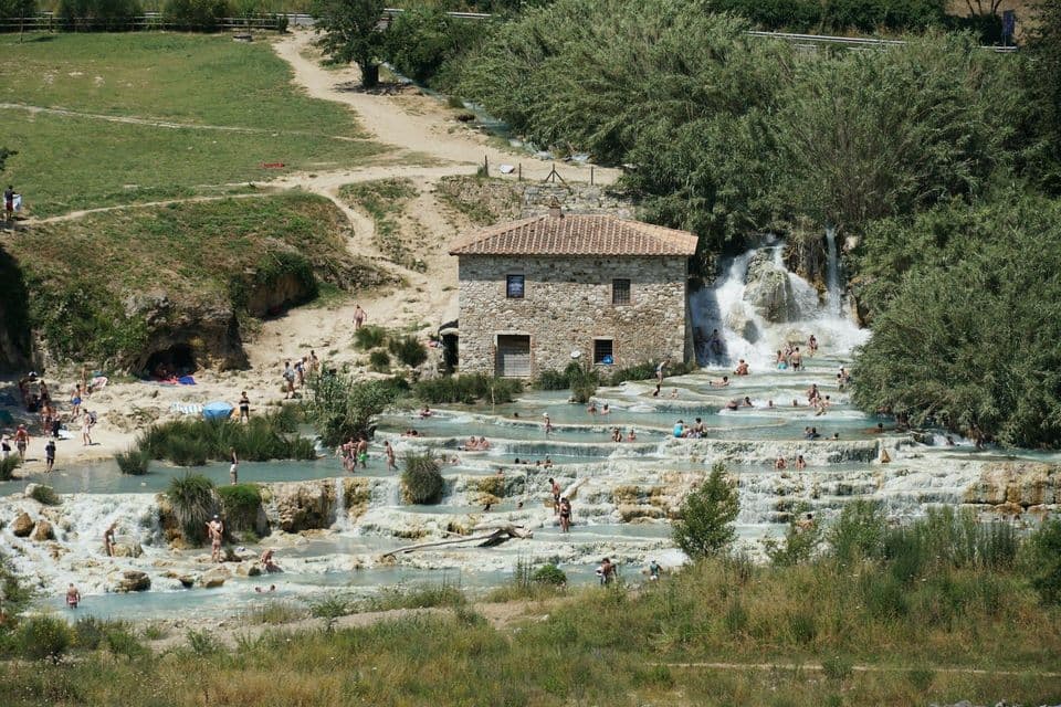Persone che fanno il bagno in piscine termali a cascata accanto a un edificio in pietra in un paesaggio lussureggiante e verde.