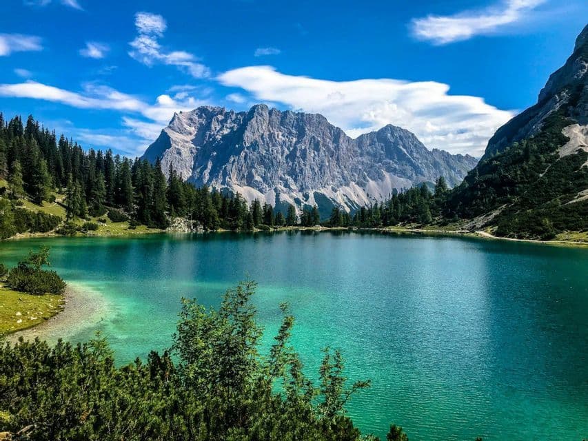 Un lago alpino turchese circondato da una pineta verde alla base di montagne rocciose sotto un cielo azzurro.