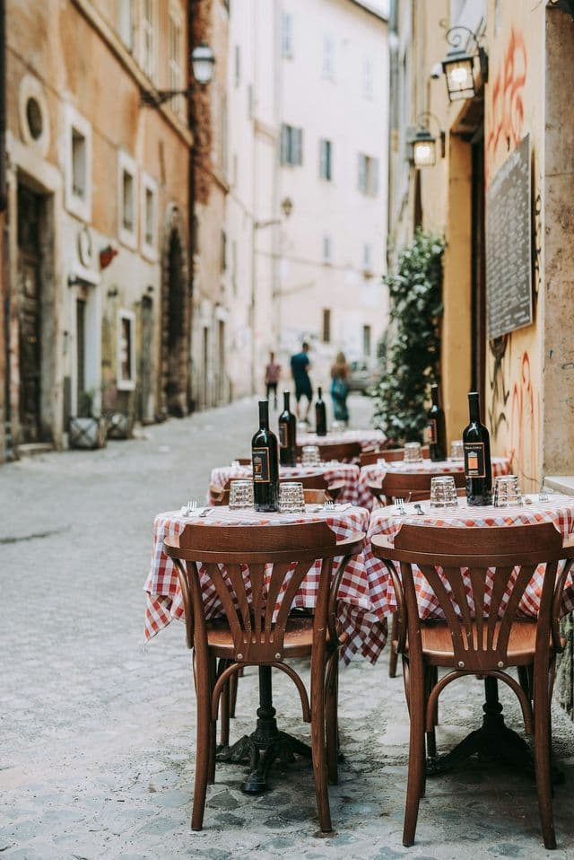 Su una stretta strada acciottolata sono disposti tavolini da caffè all'aperto con tovaglie a quadri rossi e bianchi e bottiglie di vino.