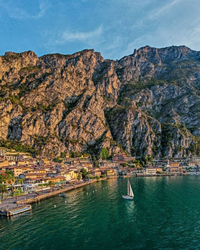 Una vista aerea di una barca a vela su acqua verde vicino a una cittadina sul lago adagiata ai piedi di una grande montagna rocciosa.