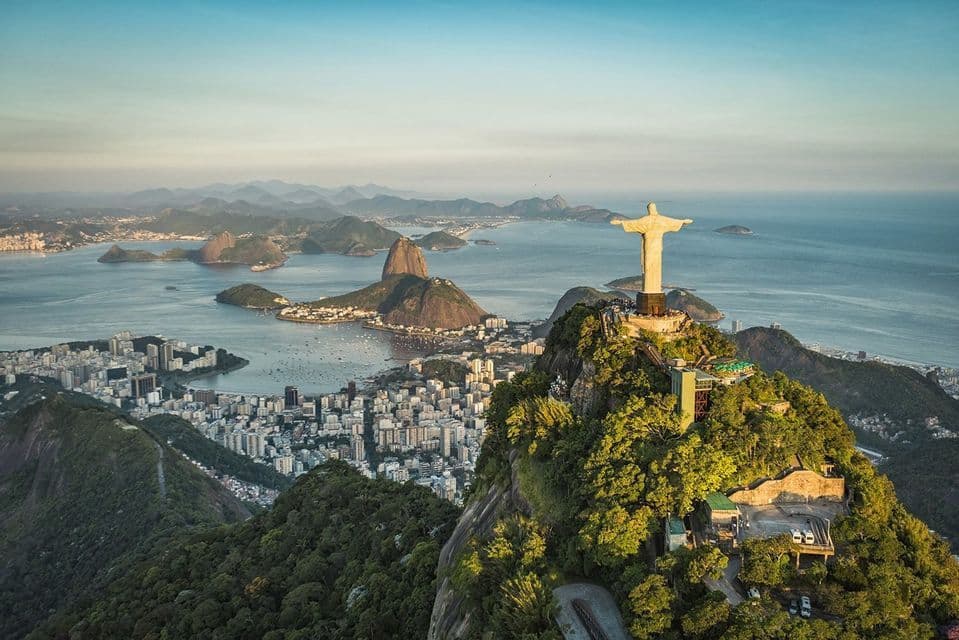 The Christ the Redeemer statue on a mountaintop overlooks a vast coastal city, a bay, and Sugarloaf Mountain at sunrise.