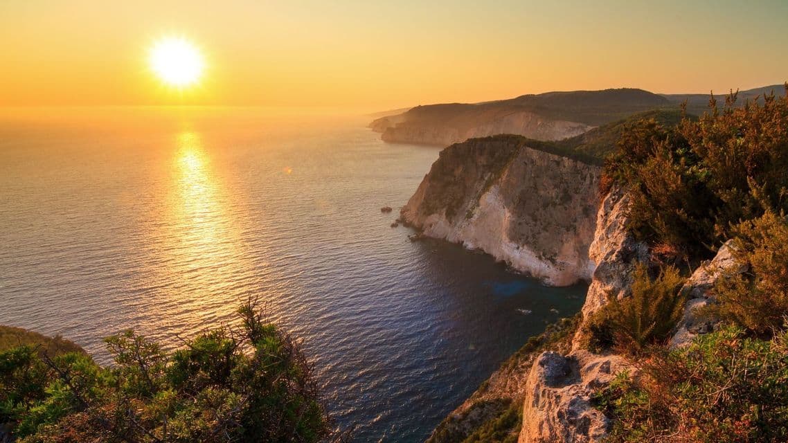 Tramonto dorato sull'oceano, visto da una scogliera che domina una costa frastagliata con vegetazione verde.
