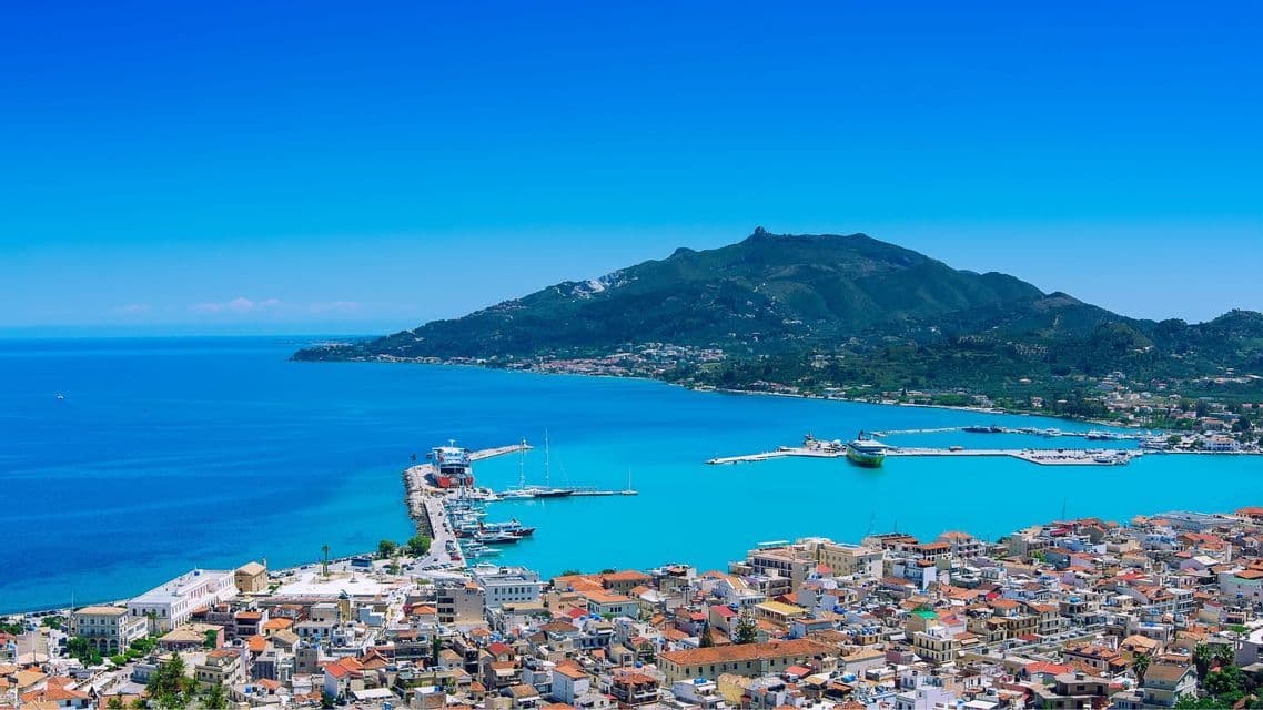 Una vista dall'alto di una città costiera con un porto su una baia turchese, con una grande montagna verde sullo sfondo.