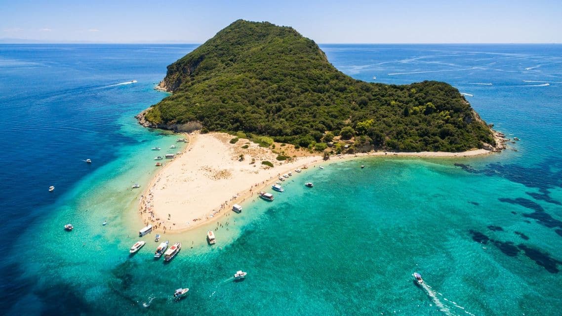 Una veduta aerea di un'isola verde e collinare con una spiaggia sabbiosa dove persone e barche si radunano nel mare turchese.