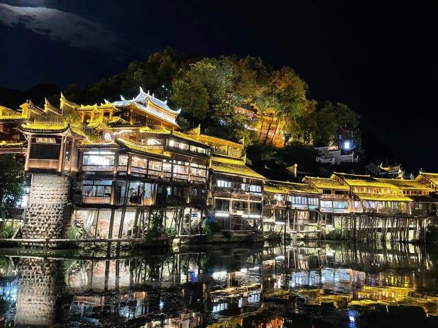 Case tradizionali su palafitte con tetti a pagoda illuminati costeggiano un fiume di notte, le cui luci si riflettono sulla superficie dell'acqua.