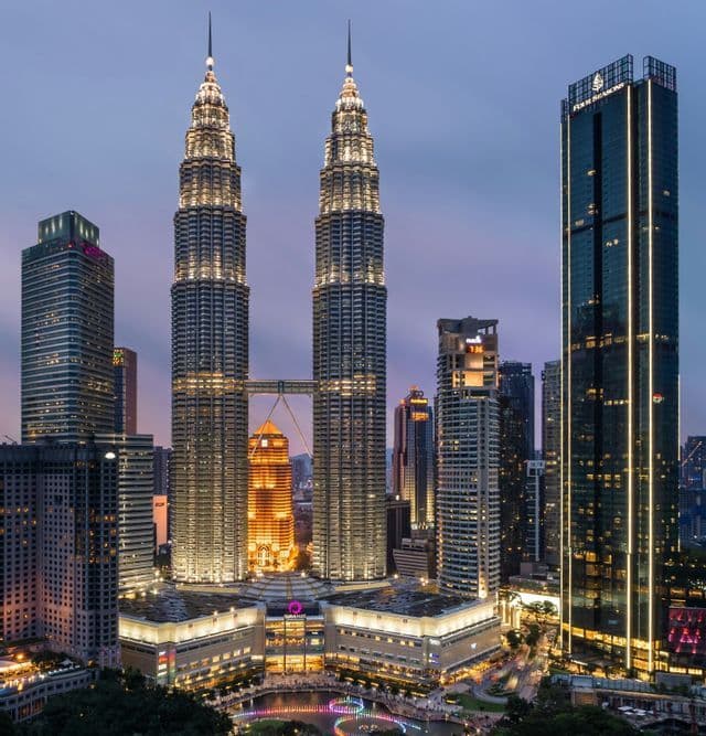 Des gratte-ciel jumeaux illuminés avec une passerelle aérienne se dressent dans un paysage urbain moderne au crépuscule.