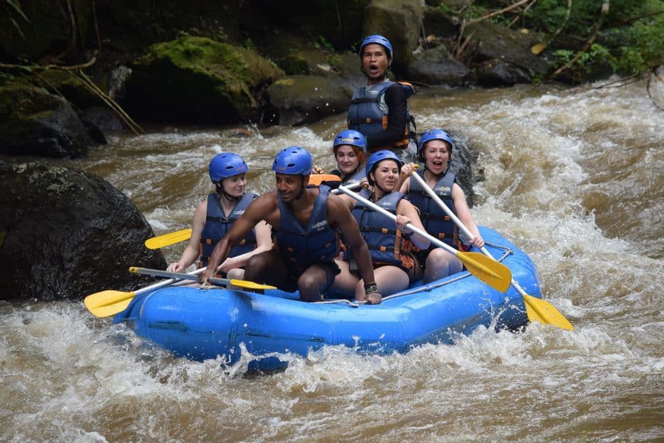 A WeRoad group trip wearing helmets and life vests paddles a blue inflatable raft through turbulent river rapids.