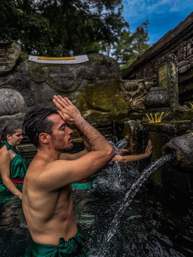 A man from a WeRoad group trip participates in a water purification ritual, praying with hands together under a stone fountain.
