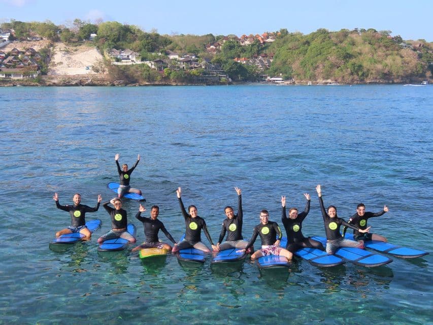 A WeRoad group trip in wetsuits sits on surfboards in the ocean, smiling and waving with a coastline behind them.