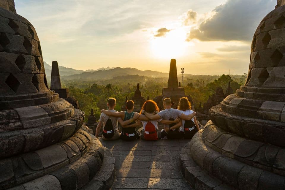 A WeRoad group trip sits with arms around each other on a stone temple, watching the sunrise over a lush landscape with stupas.