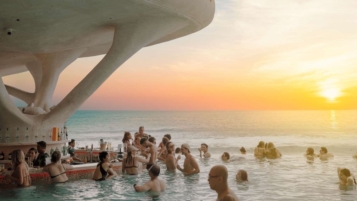 A WeRoad group trip socializing at a modern swim-up bar in an infinity pool overlooking the ocean at sunset.