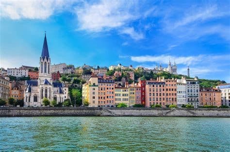 A cityscape with colorful buildings and a church with a tall steeple lining a river under a blue sky.