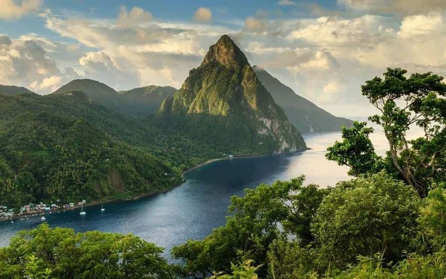 Una vista dall'alto di un'imponente montagna appuntita, ricoperta di vegetazione lussureggiante, che si protende in una baia azzurra e tranquilla con un piccolo villaggio sulla costa.