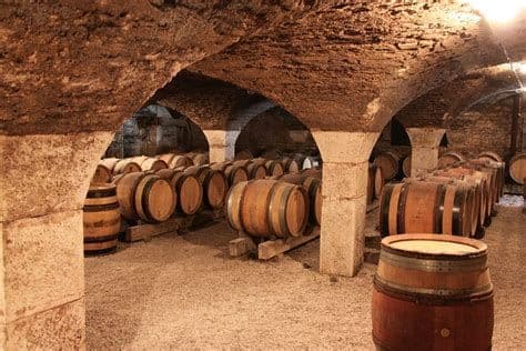 Rows of wooden barrels rest on stands in an old stone wine cellar with vaulted ceilings and thick pillars.