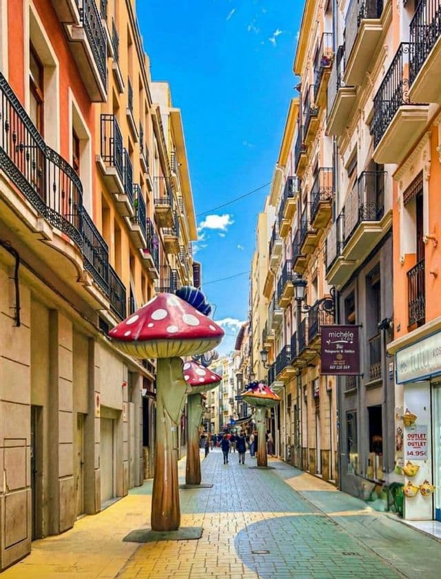 Grandi sculture di funghi rossi con macchie bianche costeggiano una stretta via pedonale tra edifici colorati a più piani.