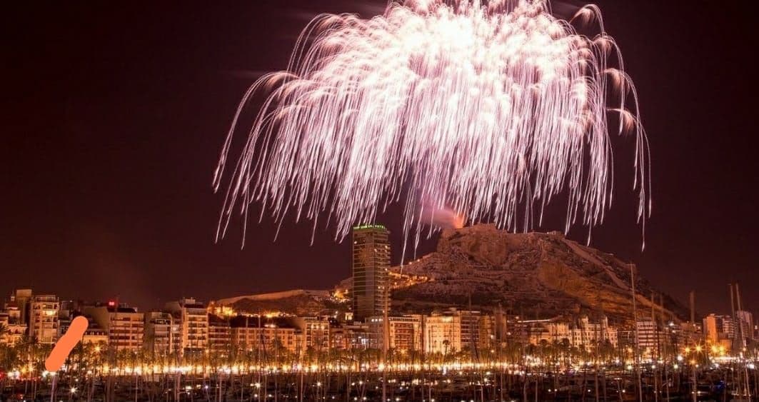 Un grande spettacolo di fuochi d'artificio esplode su una città costiera illuminata e un porto turistico di notte, con una collina sullo sfondo.