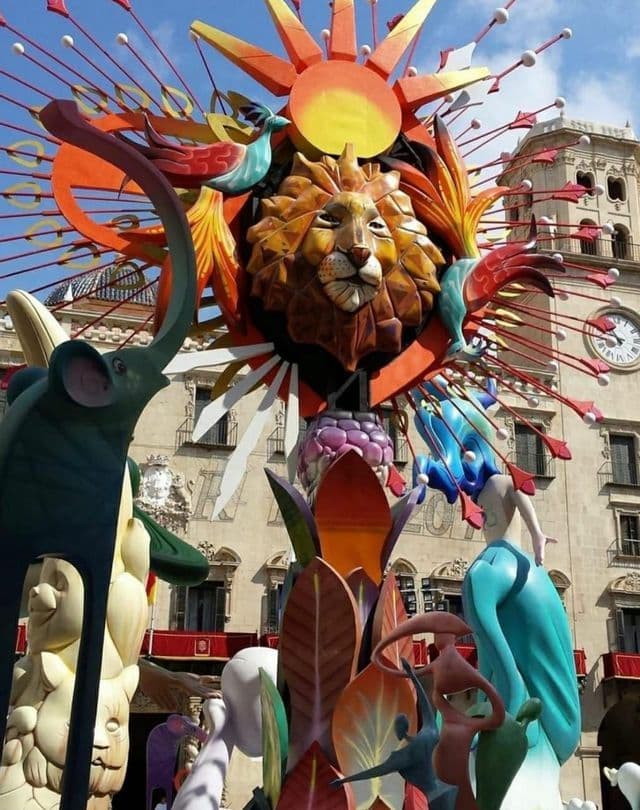 Una scultura festiva multicolore e riccamente decorata, raffigurante una testa di leone, un sole, un elefante e figure astratte, davanti a un edificio storico.