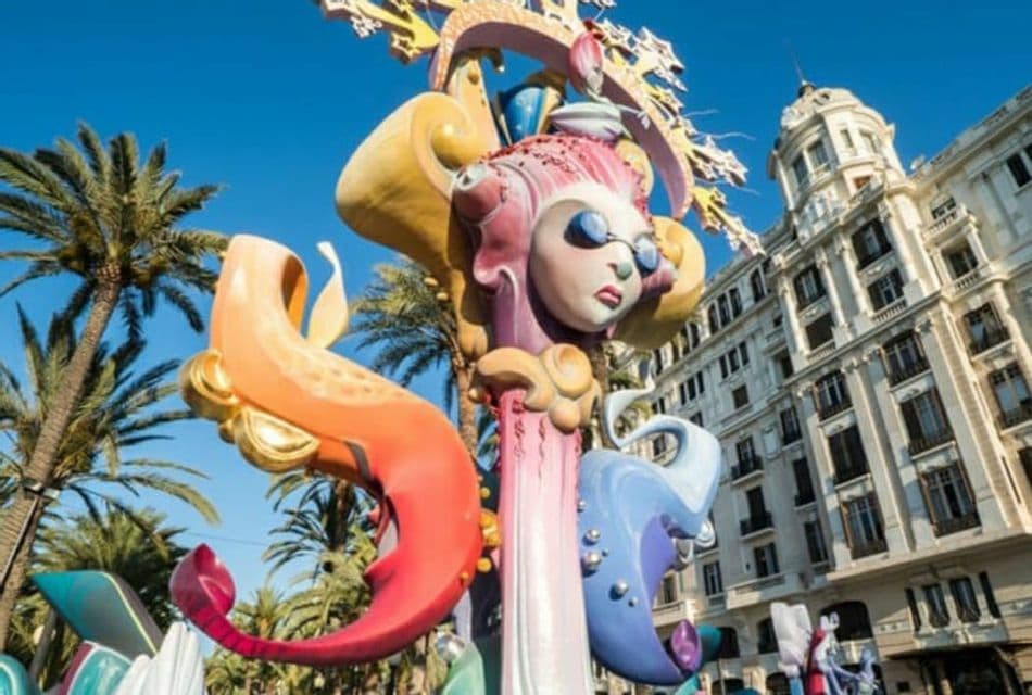 Una grande scultura colorata con un volto stilizzato si erge in una piazza cittadina con palme e un edificio decorato.