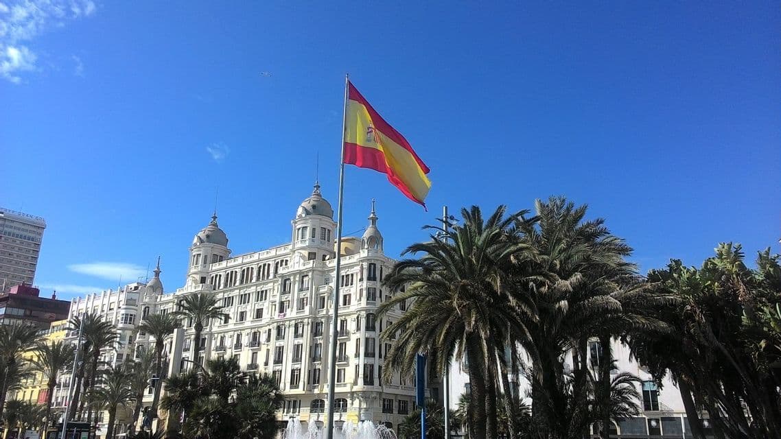 La bandiera spagnola sventola su un'asta davanti a un grande edificio bianco a più piani con palme sotto un cielo azzurro e limpido.