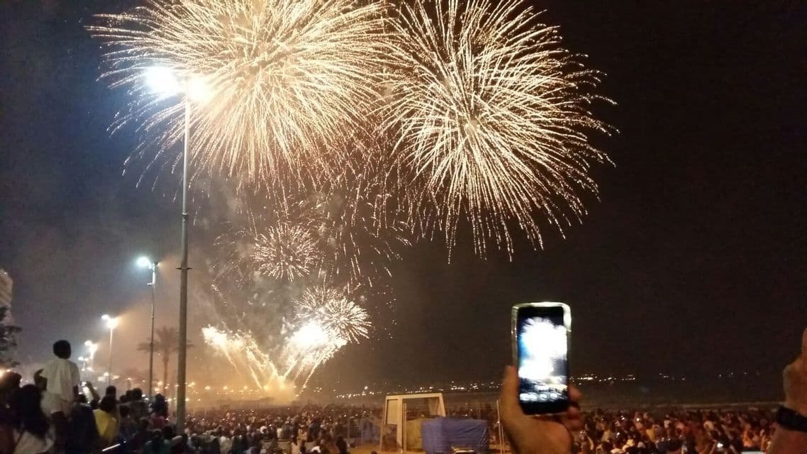 Una persona in una grande folla tiene in mano uno smartphone per registrare fuochi d'artificio dorati che esplodono nel cielo notturno sopra una spiaggia.