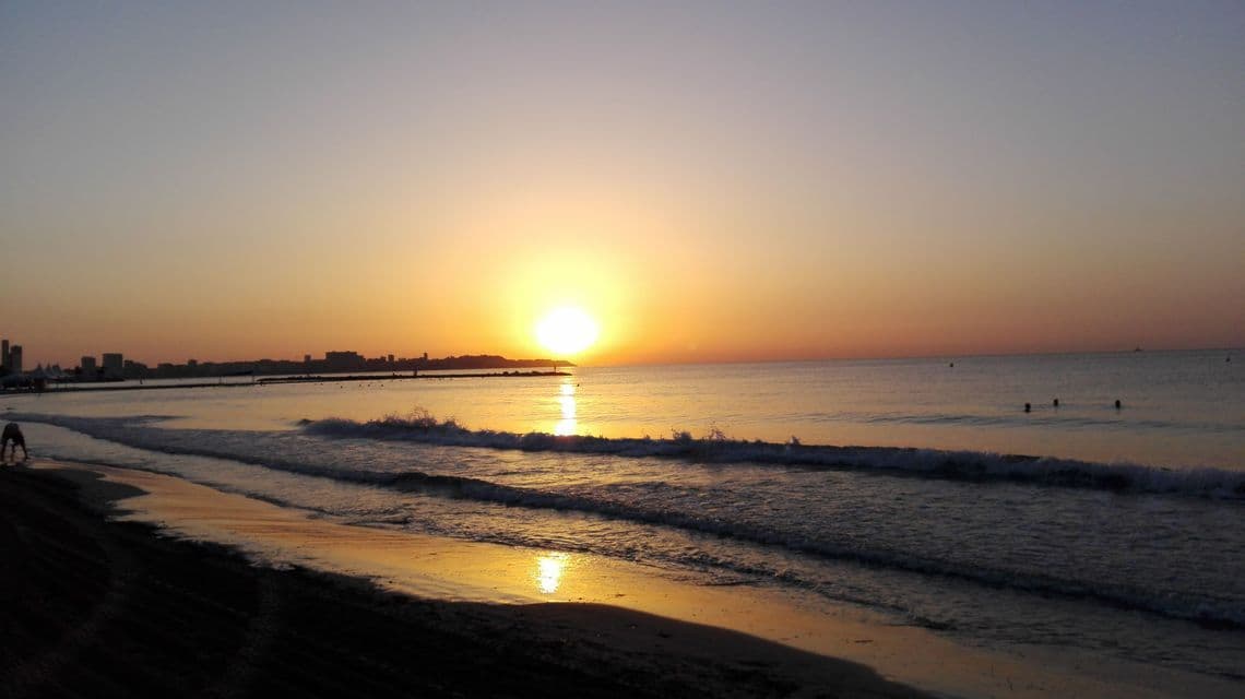 Un tramonto proietta un bagliore dorato sull'oceano mentre le onde si infrangono su una spiaggia sabbiosa, con uno skyline cittadino visibile in lontananza.