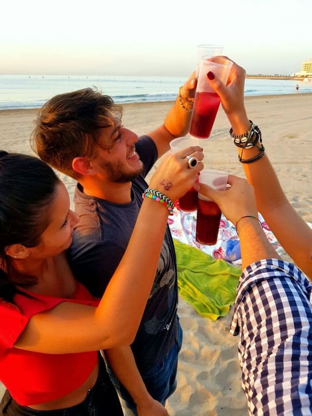 Un viaggio di gruppo WeRoad con persone sorridenti che brindano con drink in bicchieri di plastica su una spiaggia di sabbia al tramonto.