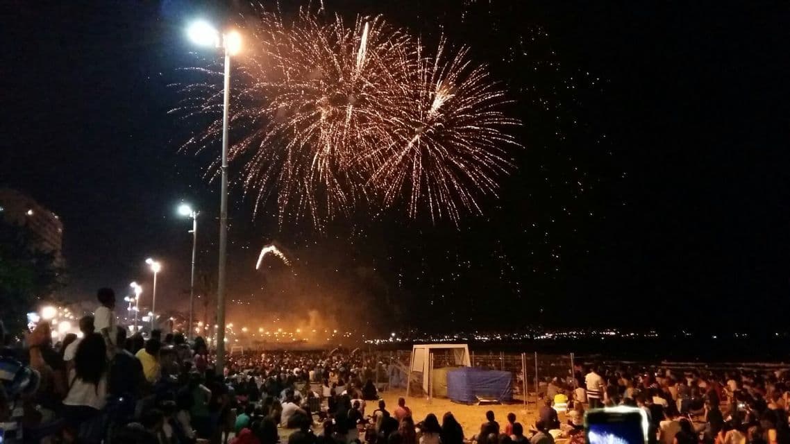 Una grande folla si è radunata su una spiaggia di notte, ammirando fuochi d'artificio dorati esplodere nel cielo scuro.