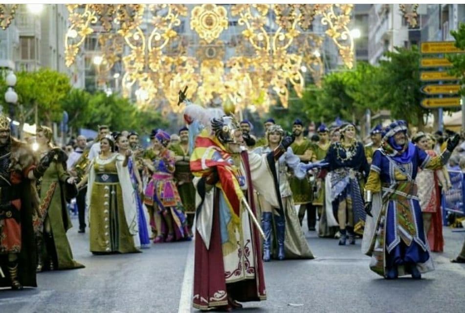 Un folto gruppo di persone in elaborati e colorati costumi sfila per le vie della città sotto sfarzose luci dorate.