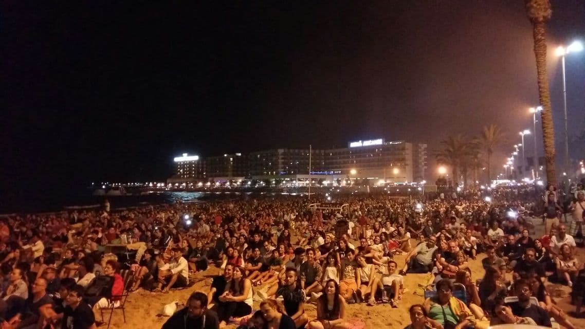Una grande folla di persone siede su una spiaggia di notte, con edifici illuminati e lampioni sullo sfondo.