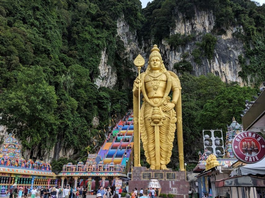 Une grande statue dorée d'une divinité se dresse au pied d'un long escalier coloré qui mène à un flanc de montagne luxuriant et rocheux.
