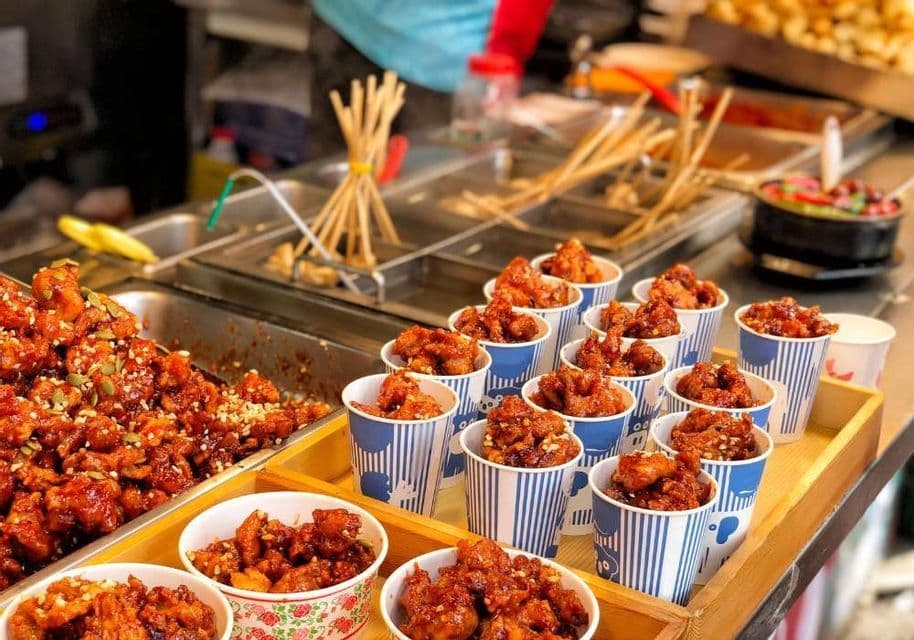 Fried chicken pieces coated in a spicy red sauce and seeds, served in striped paper cups at a street food stall.