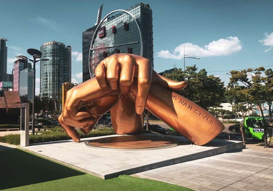 A large bronze 'Gangnam Style' sculpture of crossed hands in a modern city square with skyscrapers in the background.