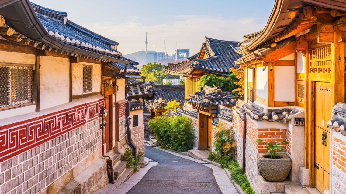 A narrow street winds through a traditional village with tiled-roof houses, with a modern city skyline in the background.