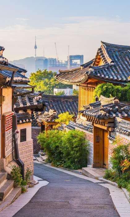 Traditional houses with tiled roofs line a narrow alley, with a modern city skyline and communications tower visible in the background.