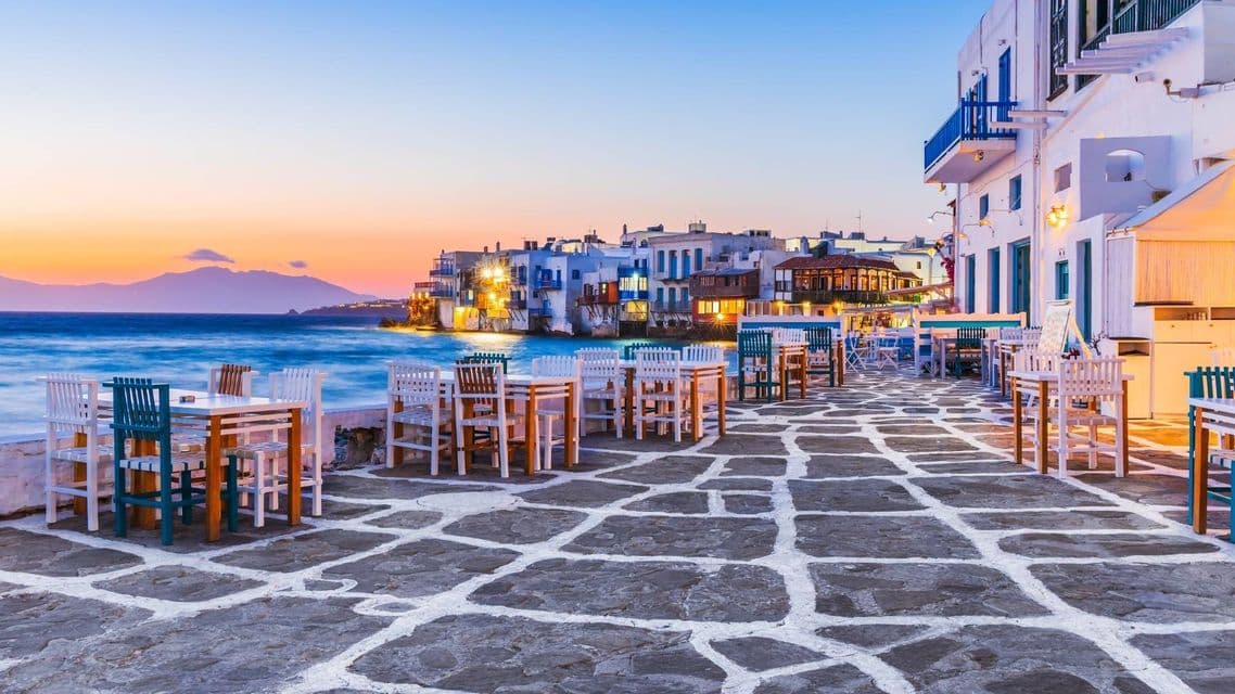 Tavoli e sedie vuoti di un bar sul lungomare in pietra al tramonto, con edifici costieri sullo sfondo.