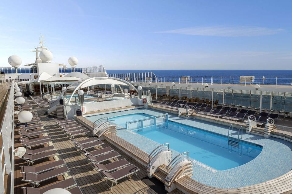 Il ponte vuoto di una nave da crociera con piscina e sedie a sdraio che si affaccia sull'oceano blu in una giornata di sole.